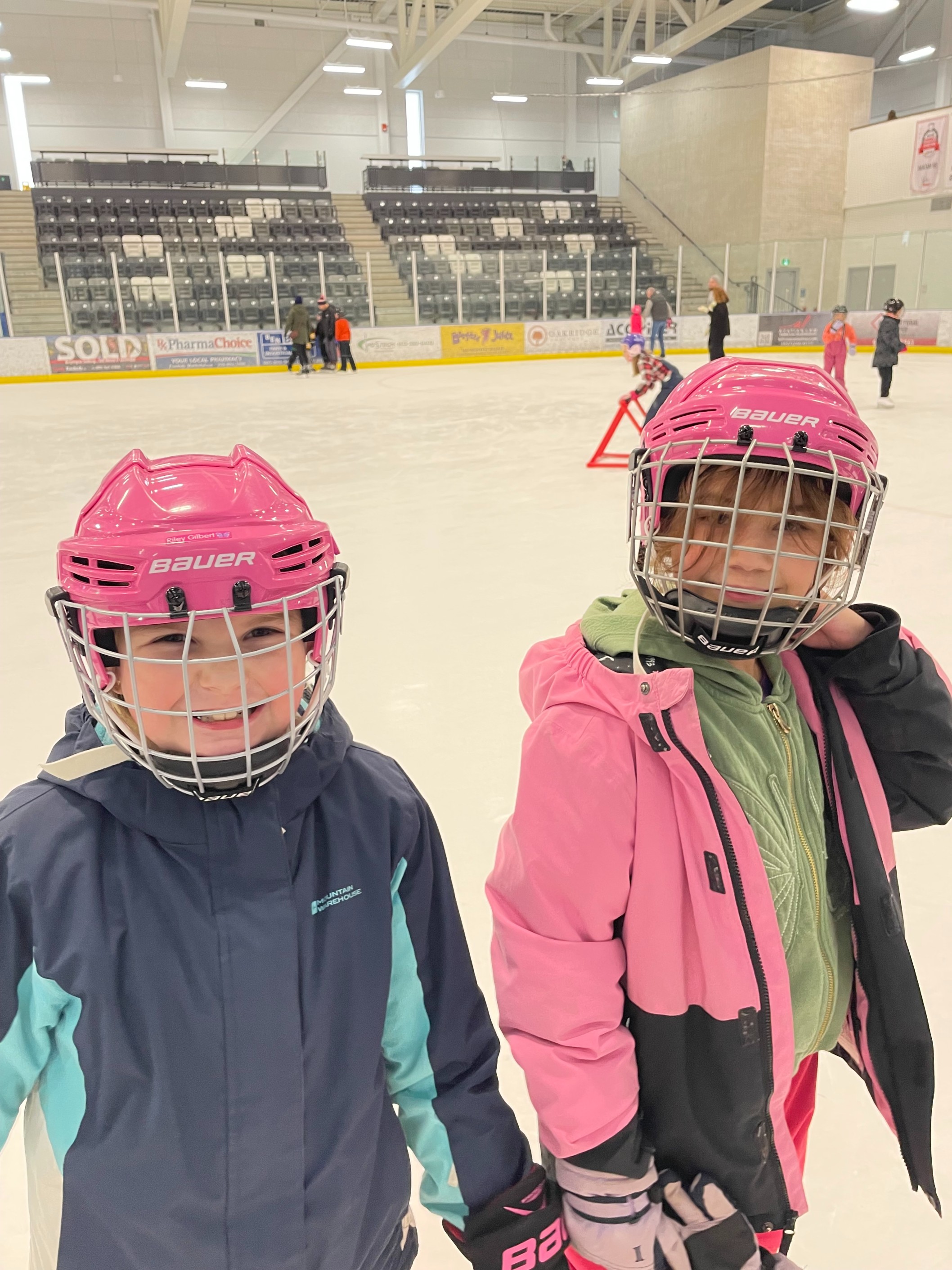 Two École élémentaire Nouvel Horizon students skating together on an ice rink wearing pink helmets and winter jackets.